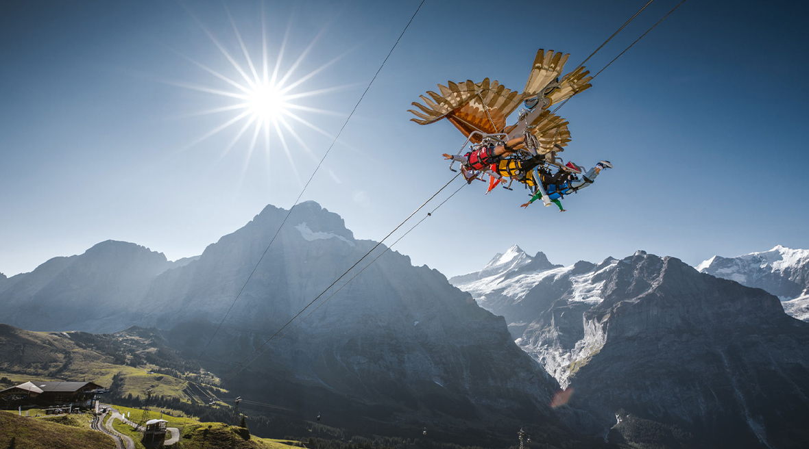 A group zip wire descends the Grindelwald First mountain. The flyers are lying face down and the zip is decorated like a big bird